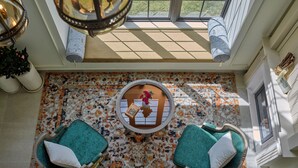 A sunlit room with a patterned floor, two teal cushioned chairs, a small table with a vase of flowers, and a large window with a view of greenery. - Grand Mercure Dalat (Lam Vien - Da Lat)