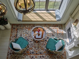 Une pièce baignée de lumière, avec un sol à motifs, deux fauteuils turquoise, une petite table ornée d’un vase de fleurs et une grande fenêtre donnant sur un paysage verdoyant.