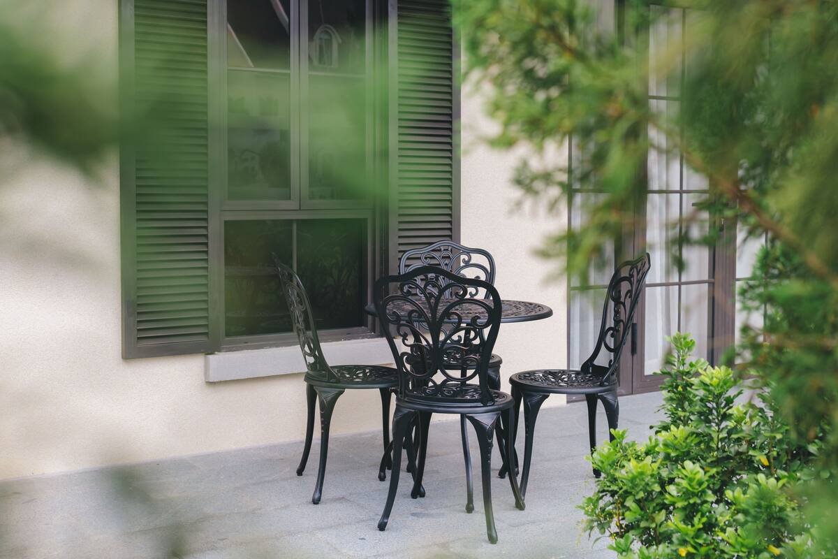 A patio with a round table and four chairs, a window with green shutters, and a small green bush.
