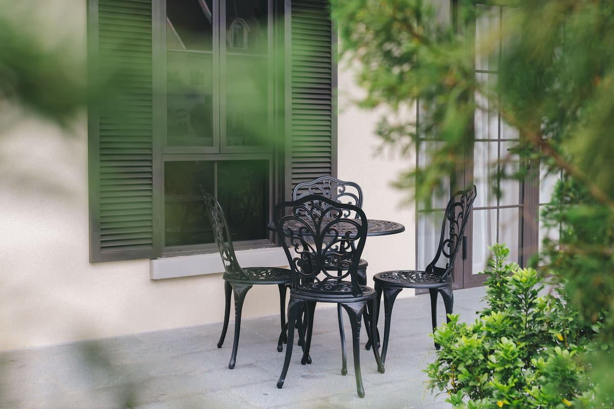 A patio with a round table and four chairs, a window with green shutters, and a small green bush.