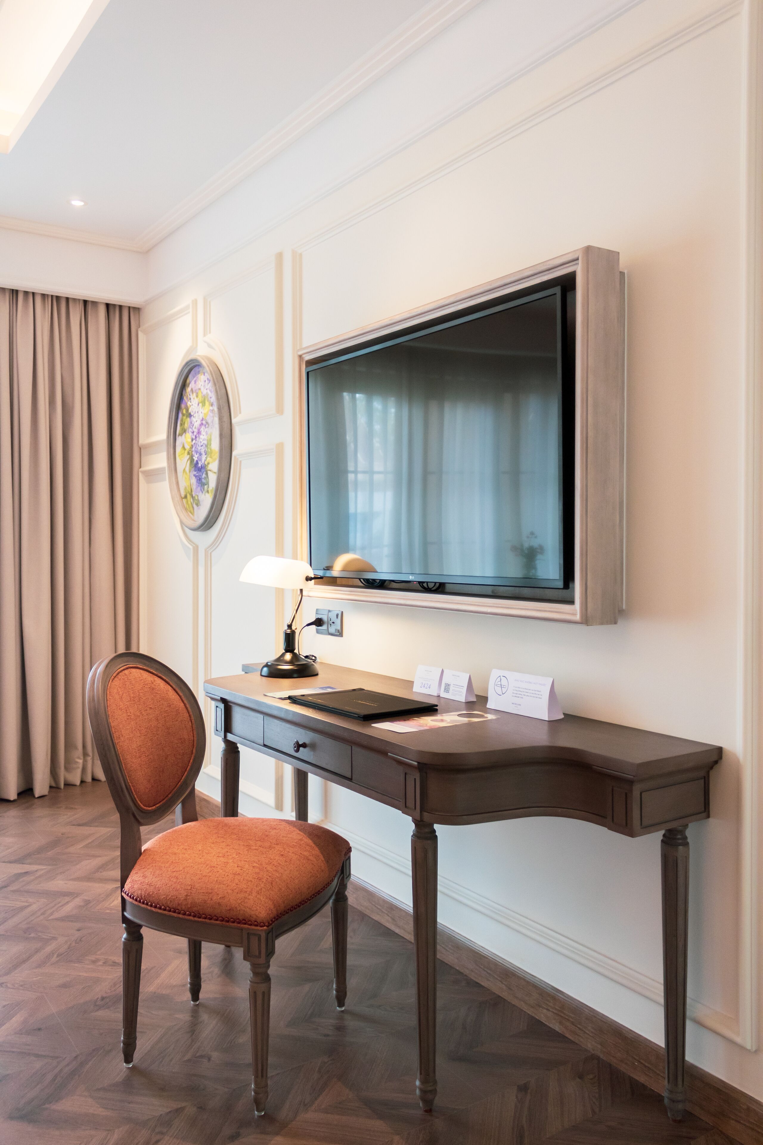 A hotel room with a wooden desk, a chair, a flat-screen TV, a lamp, and a framed picture on the wall.