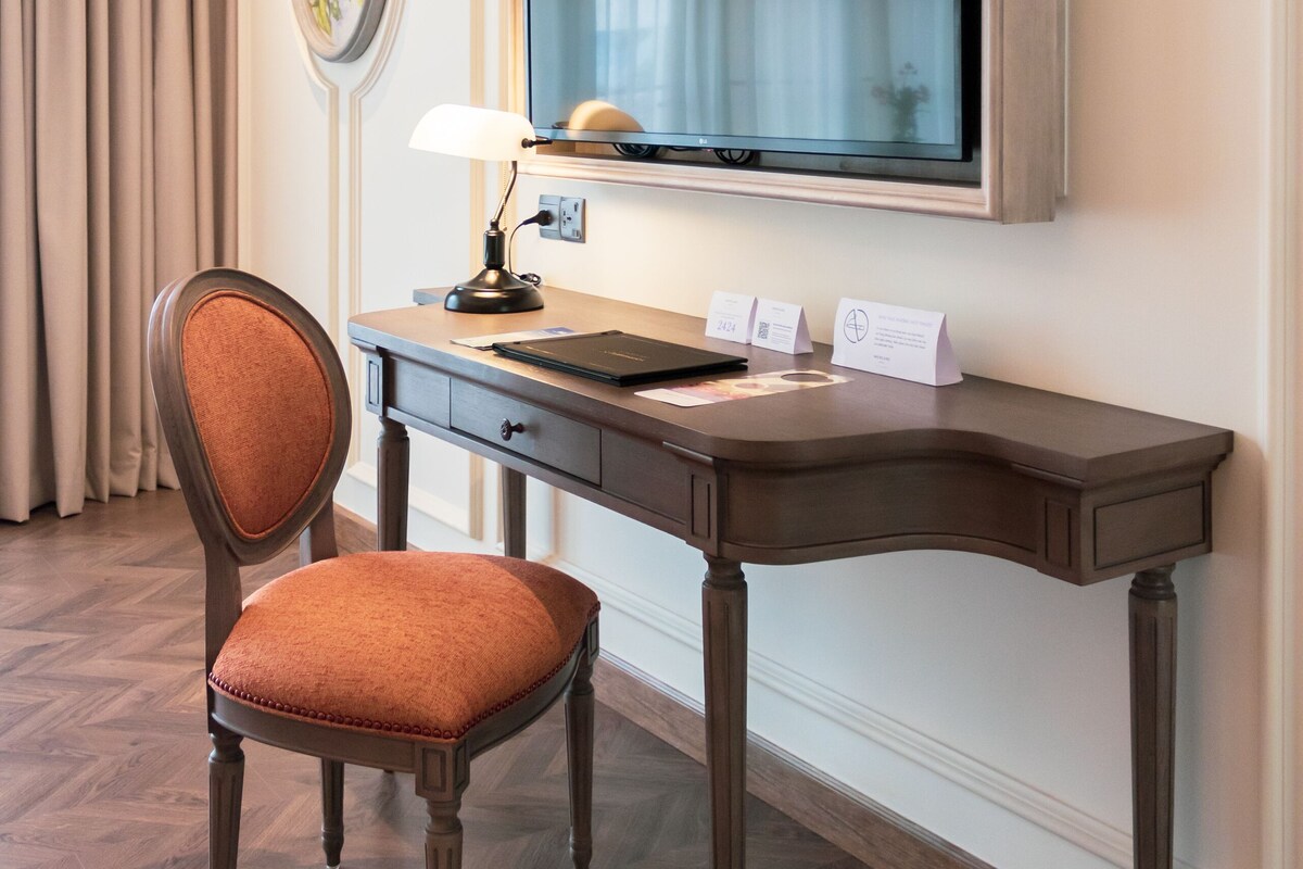 A hotel room with a wooden desk, a chair, a flat-screen TV, a lamp, and a framed picture on the wall.