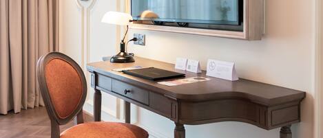 A hotel room with a wooden desk, a chair, a flat-screen TV, a lamp, and a framed picture on the wall.