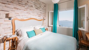 A bedroom with a wooden headboard, a bed with white and blue bedding, a nightstand with a phone, a wooden chair, and a view of the sea through a window with teal curtains. - Ikion Eco Boutique Hotel (Alonissos)