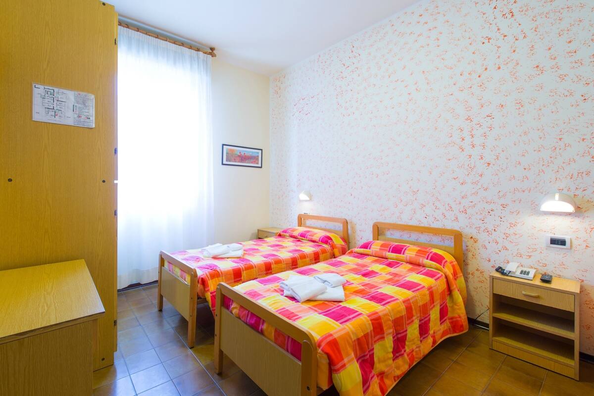 A room with two wooden beds, a wooden desk, a small bedside table, and a wall-mounted lamp.