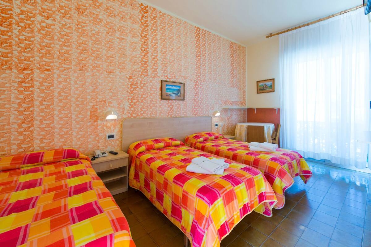A hotel room with three beds, a large window with sheer curtains, and a patterned wall.