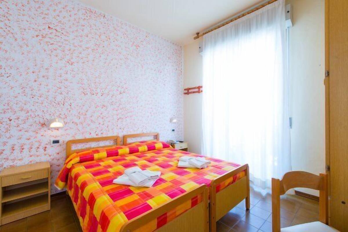 A bedroom with a floral wallpaper, a bed with a colorful quilt, two bedside tables, and a chair.