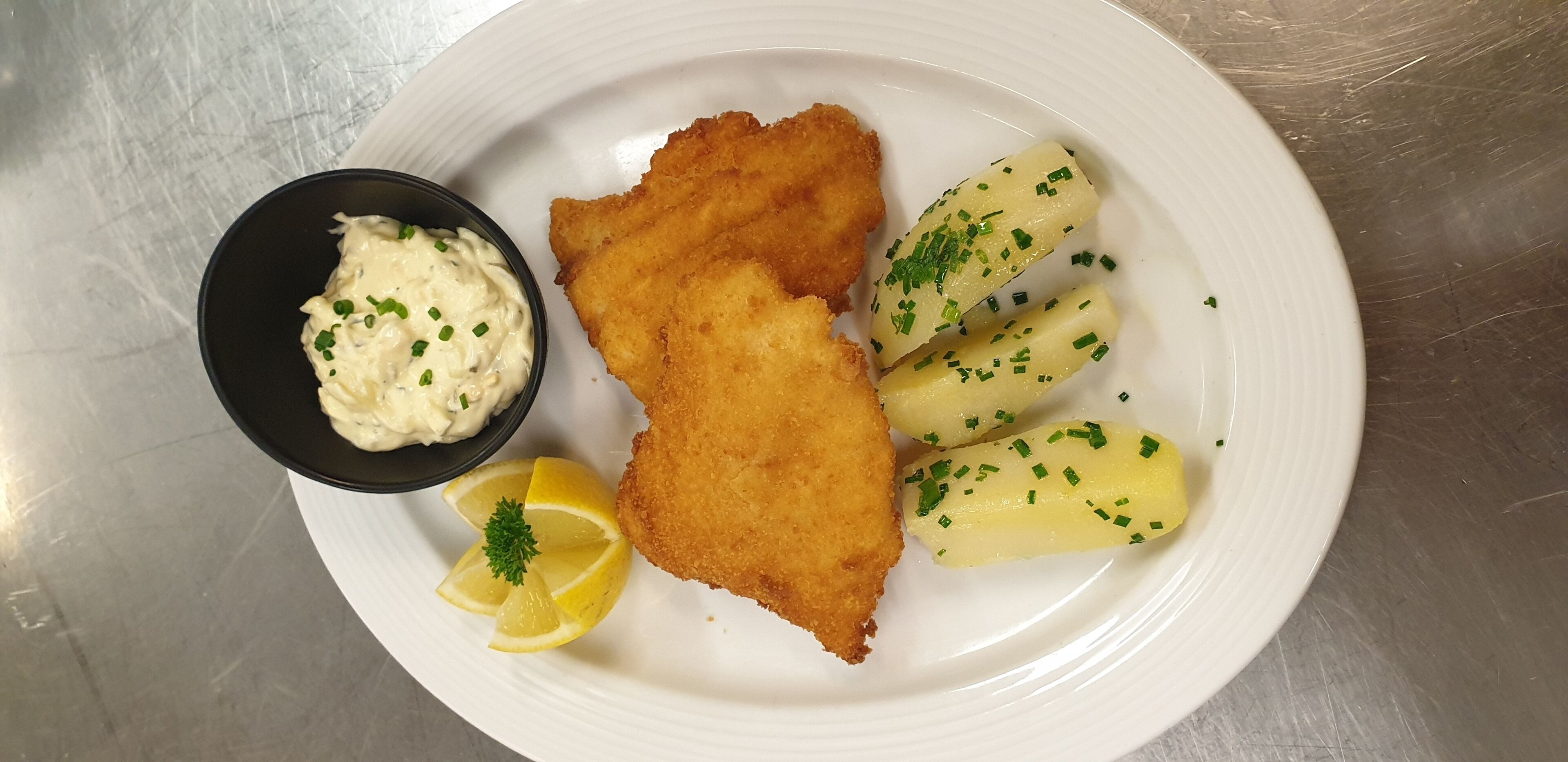 A plate with breaded fish fillets, boiled potatoes, a small bowl of sauce, and lemon slices.
