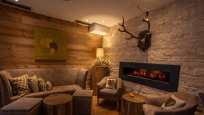 Un rincón lounge acogedor con un sofá modular, sillones y mesas redondas de madera. Se ve una chimenea con un fondo de pared de piedra y una decoración de astas de ciervo.