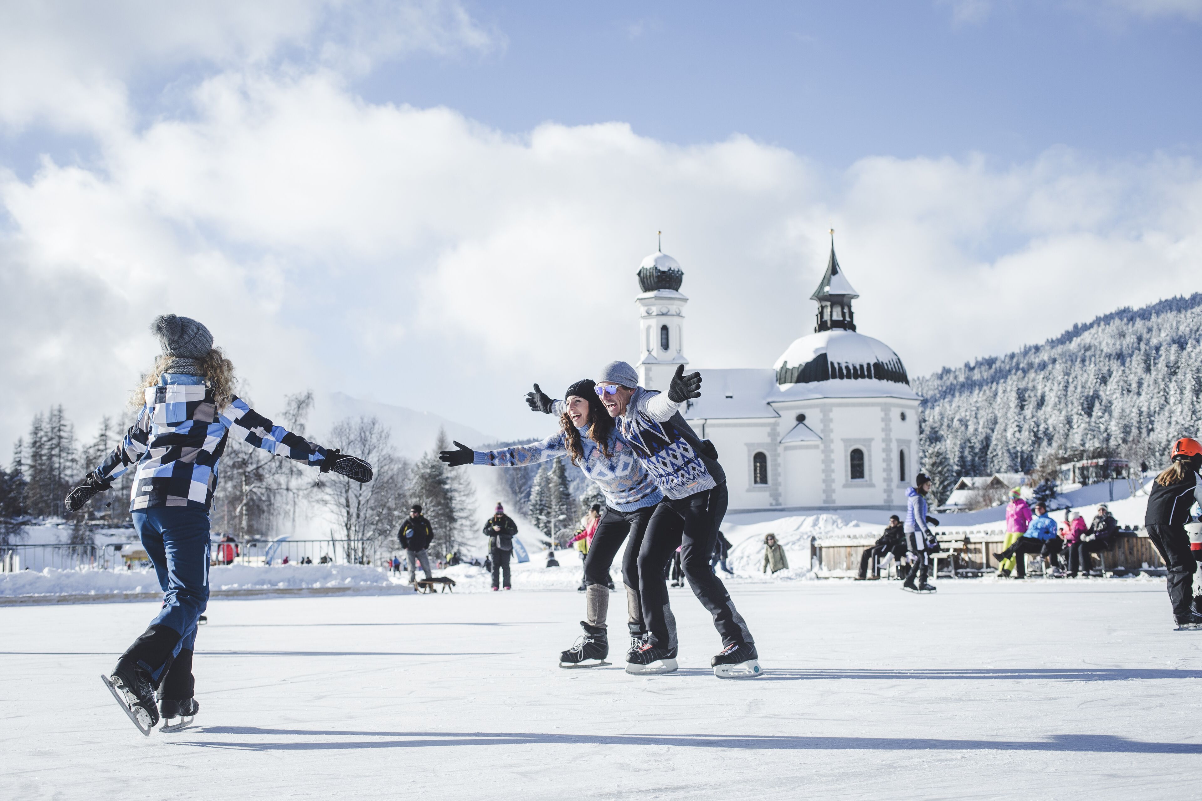 Um grupo de pessoas patinando no gelo em um lago congelado, com uma igreja e árvores cobertas de neve ao fundo.