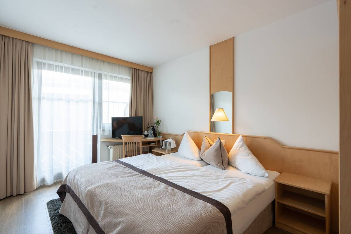Habitación de hotel con una cama grande, un escritorio con televisor, una lámpara de noche y una ventana con cortinas.