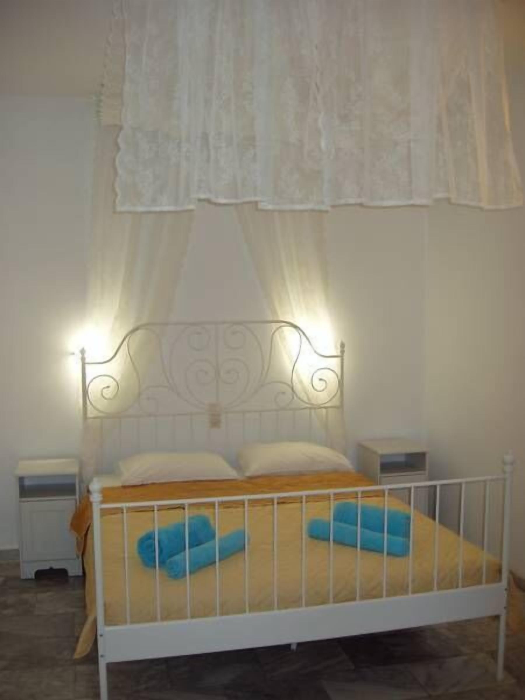 Ein Einzelbett mit weißem Metallgestell, einem Holz-Kopfstück und einem gelben Bettbezug. Zwei weiße Nachttische mit Lampen sowie ein weißer Spitzenvorhang über dem Bett.