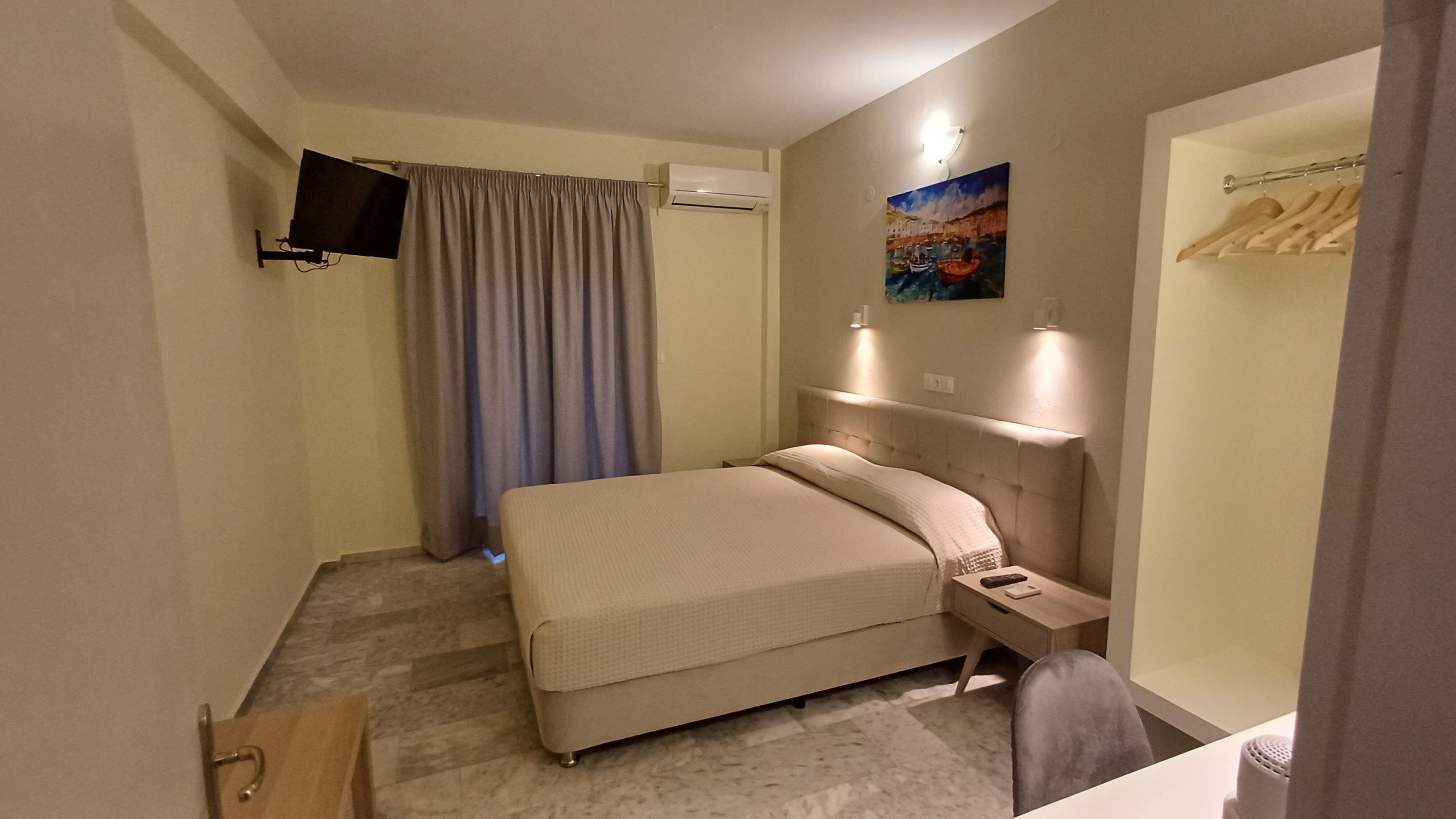 A hotel room with a bed, a TV mounted on the wall, a painting, a wardrobe, and a desk with a chair.
