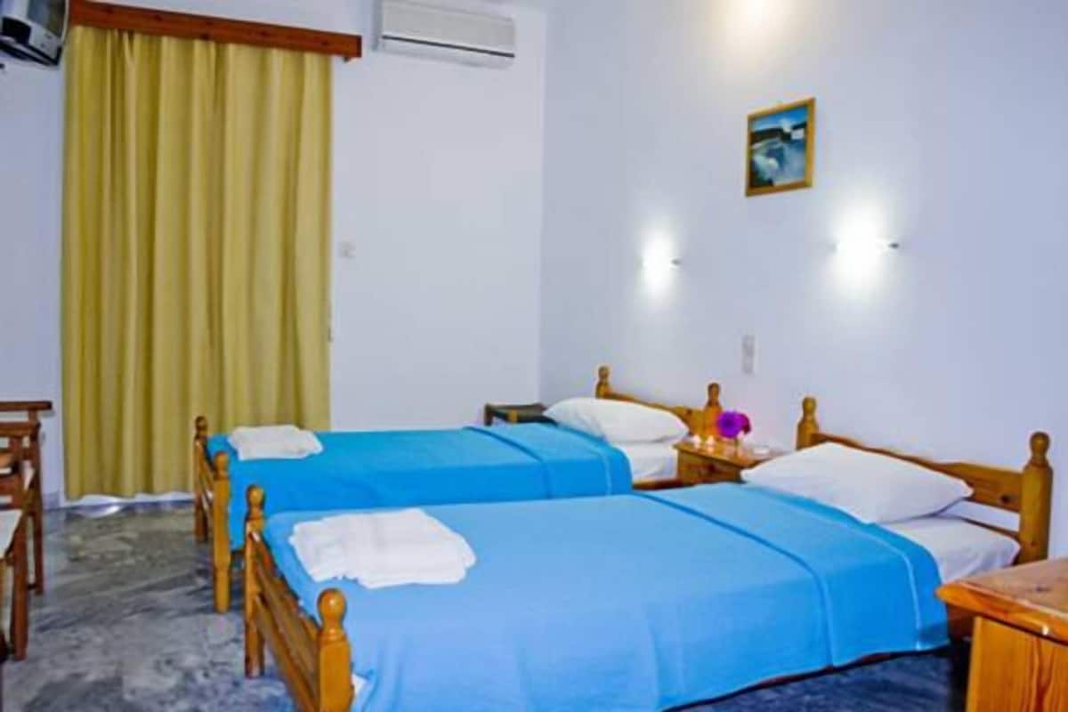 Une chambre d’hôtel avec deux lits simples, des têtes de lit en bois, une petite table de chevet avec une lampe, un climatiseur fixé au mur et une fenêtre avec des rideaux jaunes.
