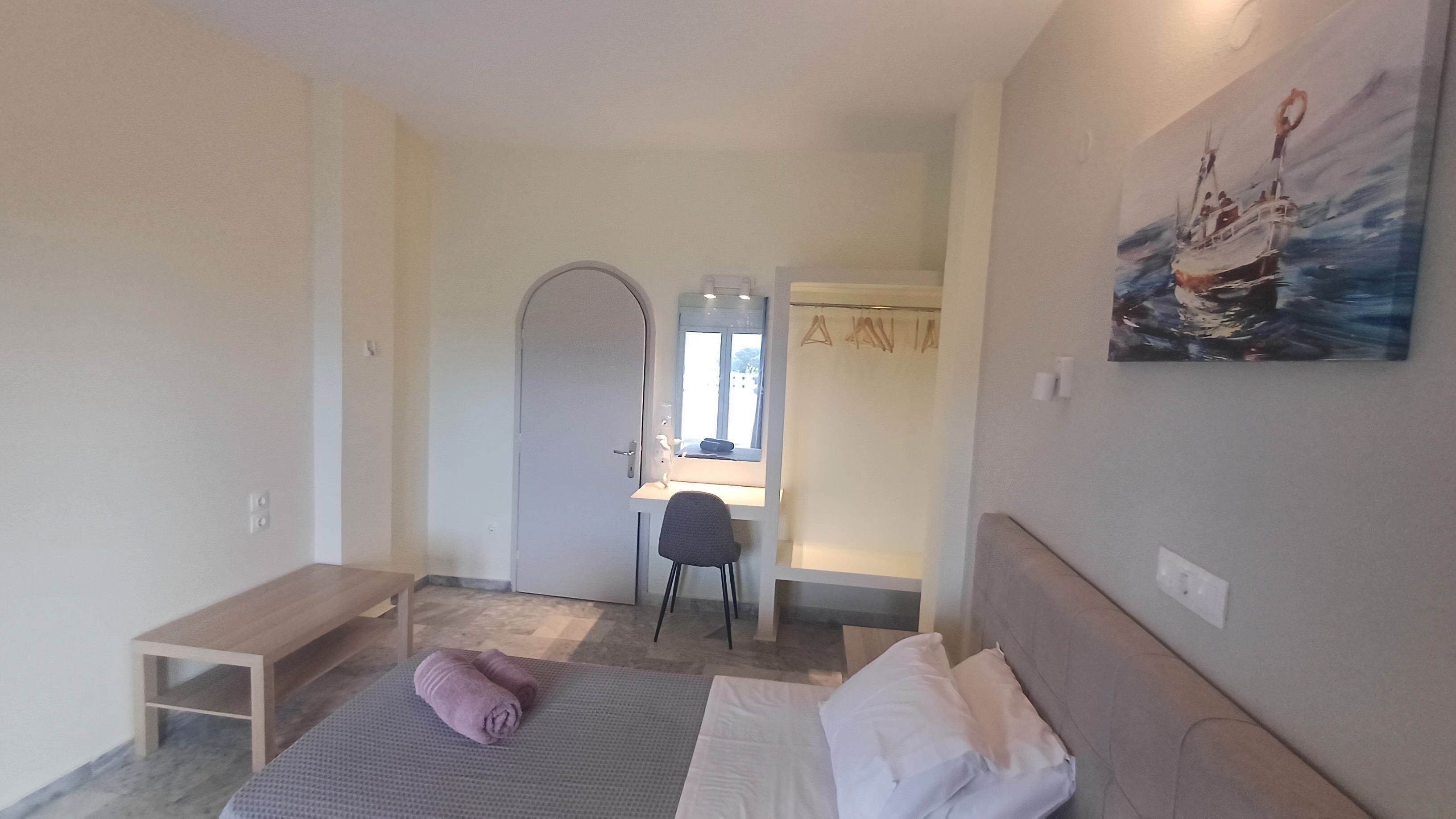 Une chambre à coucher avec un lit, un bureau et une chaise. Il y a un tableau accroché au mur et une porte qui donne sur une autre pièce.