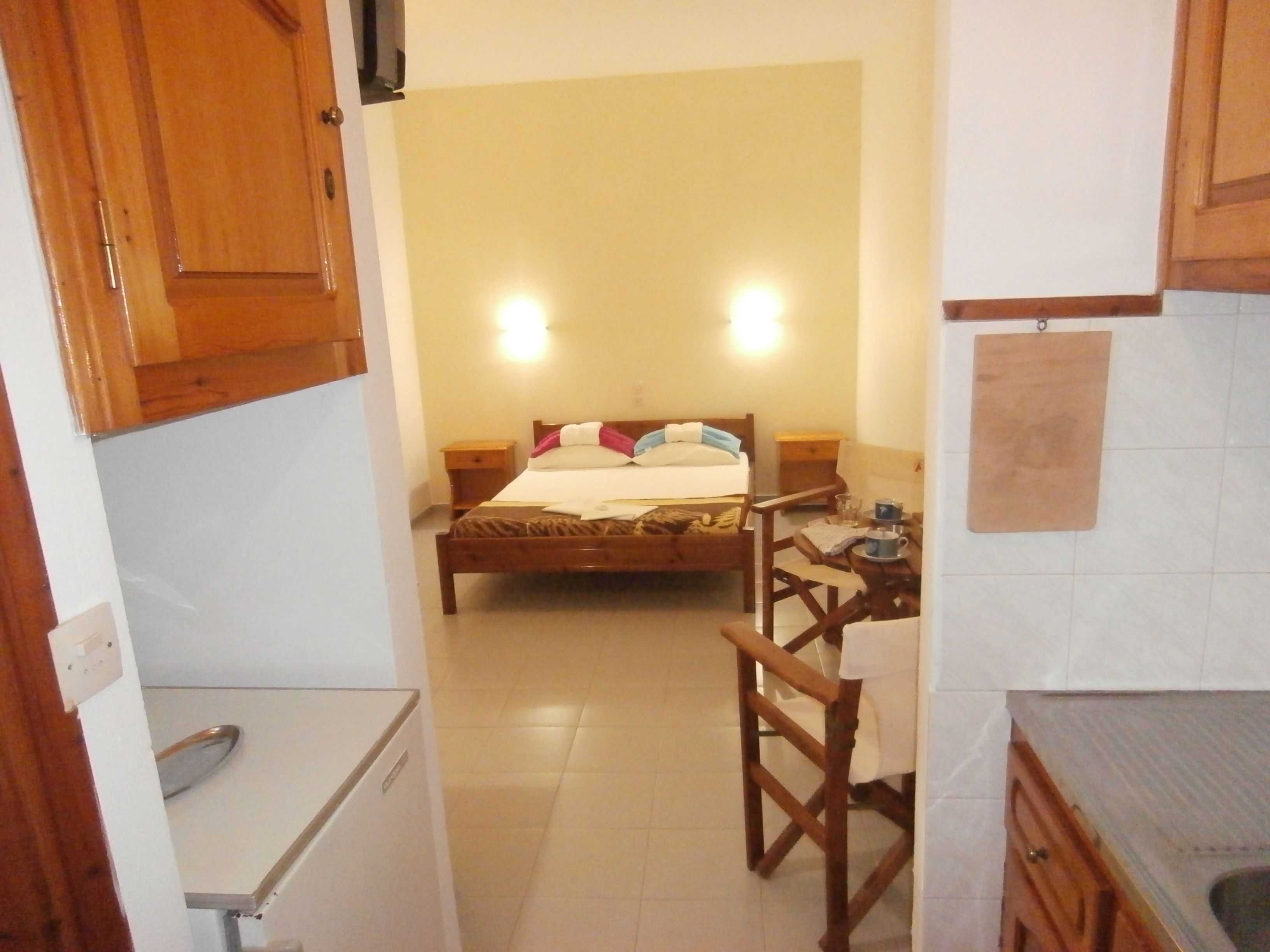 Habitación de hotel compacta con cama, una mesita con silla, kitchenette y armarios de madera.