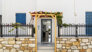 A stone-walled entrance with a wooden archway adorned with flowers, leading to a building with blue shutters and a stone facade. - Ikaros Studios & Apartments (Naxos)