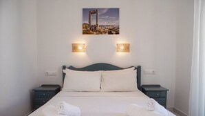 A neatly made bed with white linens and rolled towels, a dark blue headboard, and two bedside tables with wall-mounted lamps. - Ikaros Studios & Apartments (Naxos)
