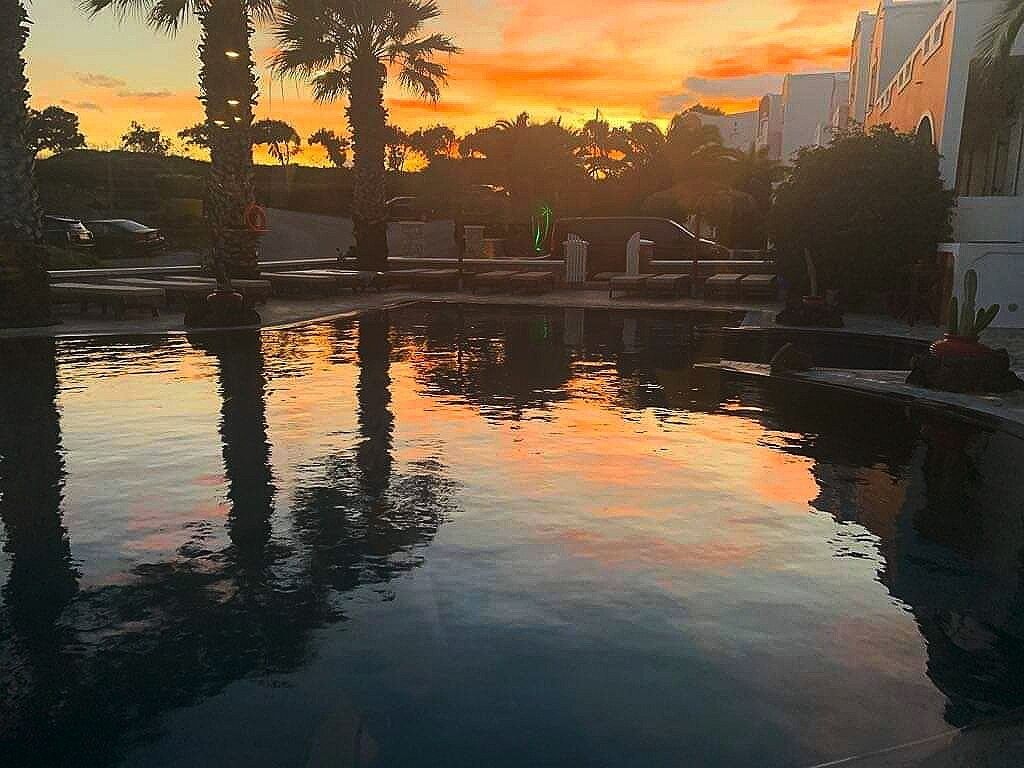 Un área de piscina tranquila con palmeras y un atardecer al fondo.