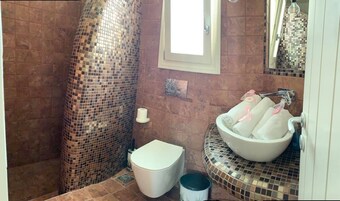Un baño con ducha revestida de mosaicos, inodoro blanco y lavabo con recipiente y dos toallas.