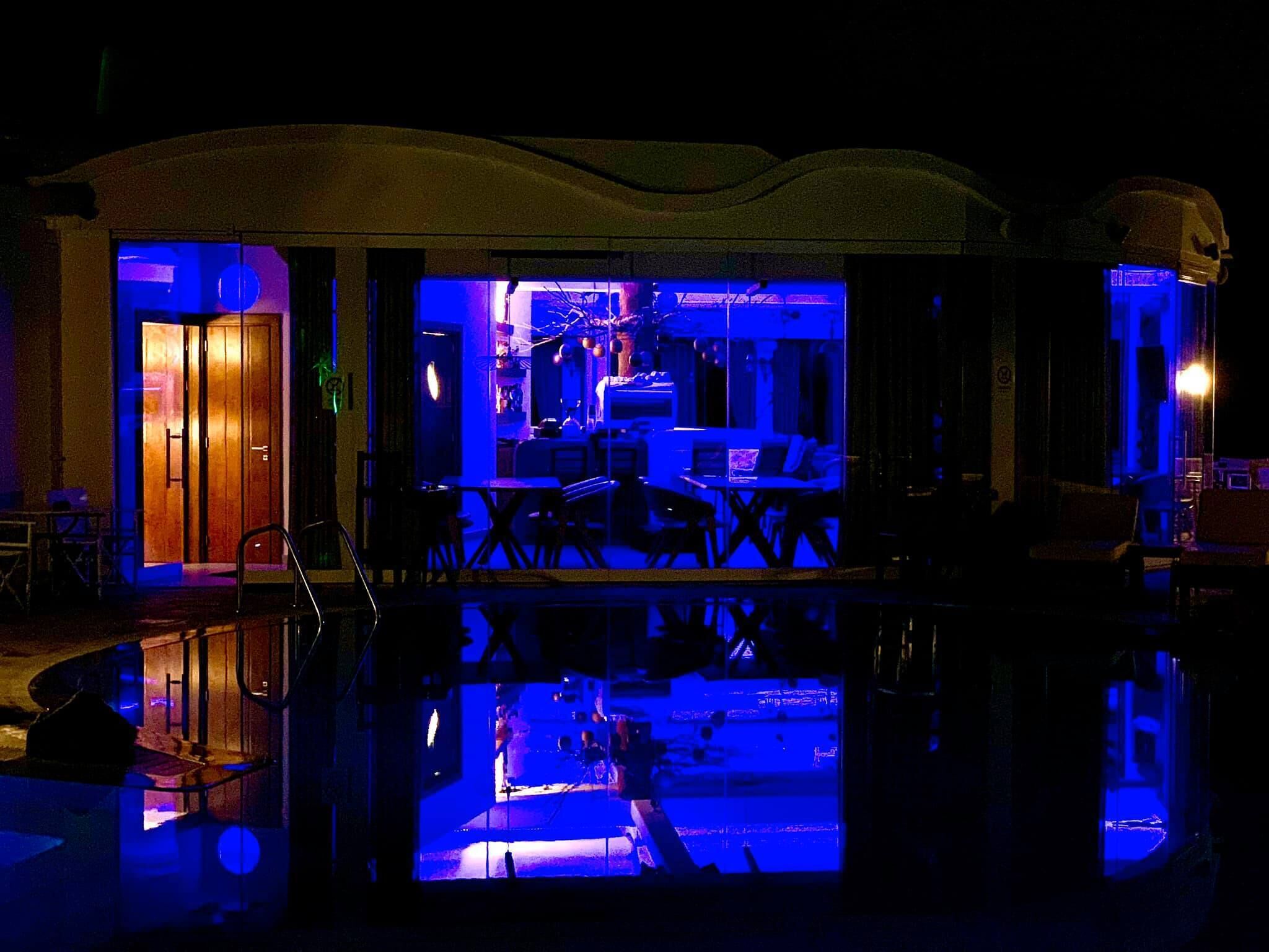 Un edificio con una piscina reflectante en primer plano, una zona de bar con iluminación azul y una puerta que da a una habitación.