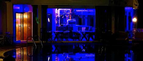 Un edificio con una piscina reflectante en primer plano, una zona de bar con iluminación azul y una puerta que da a una habitación.