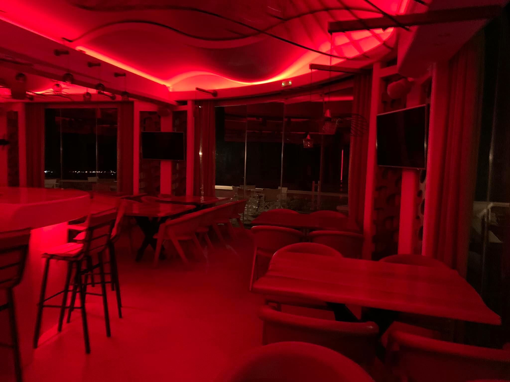 A dimly lit room with red ambient lighting, featuring a bar area with stools and tables, and large windows offering a view of the outside.