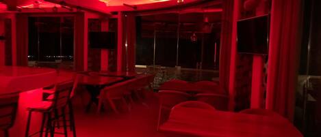 A dimly lit room with red ambient lighting, featuring a bar area with stools and tables, and large windows offering a view of the outside.