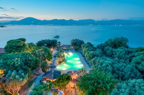 Vista aérea de un complejo turístico con piscina, rodeado de exuberante vegetación y con vistas a una masa de agua tranquila con montañas a lo lejos.
