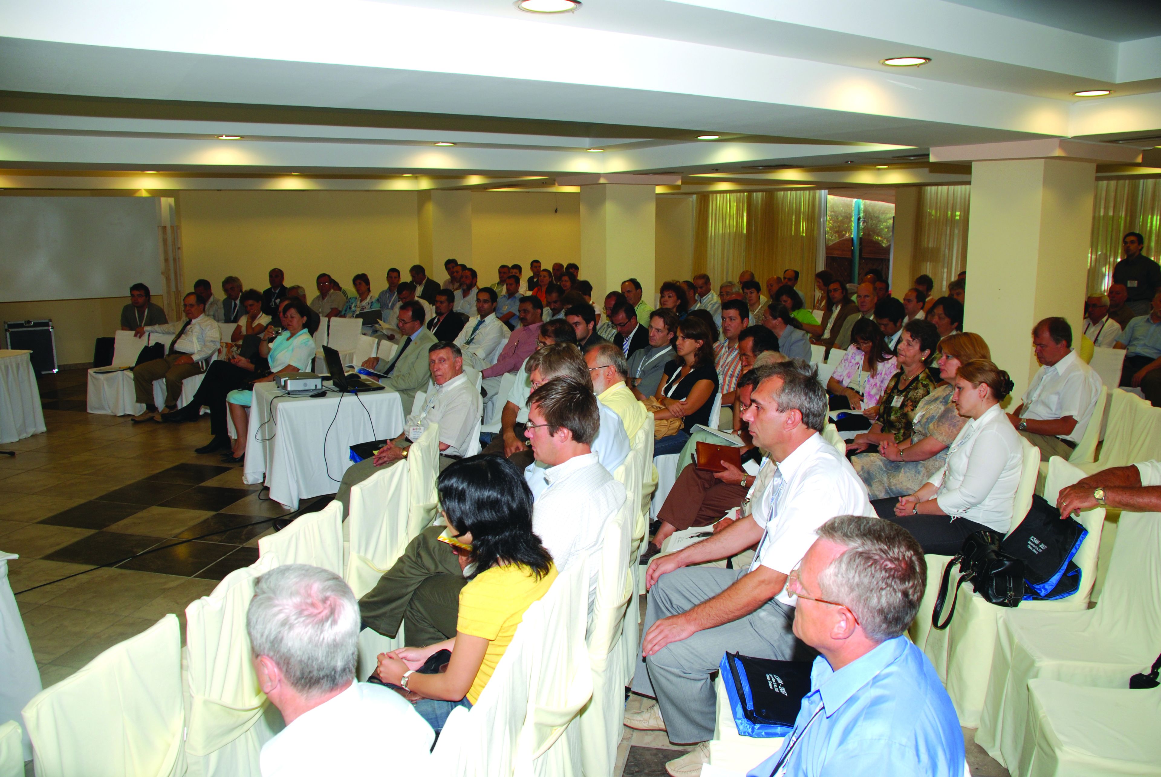 A conference room with rows of chairs and tables, a projector screen, and attendees seated and listening.