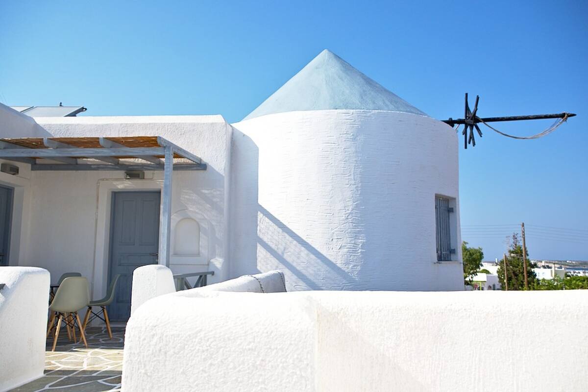 A white building with a conical roof, a door, and a window. There are chairs and a table outside.