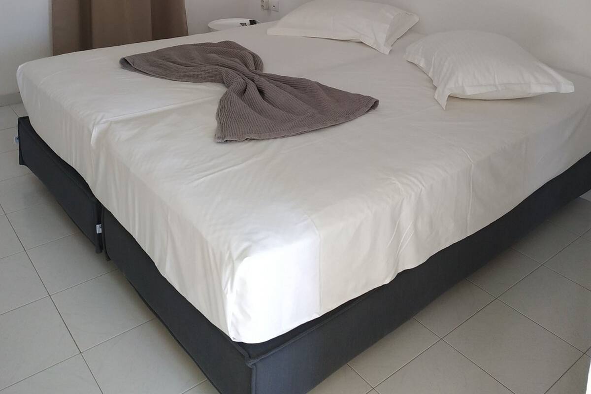 A single bed with a white mattress and dark frame, a bedside table with a lamp, and a curtain on the window.