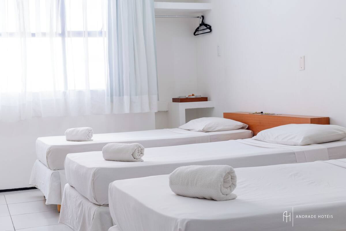 Quarto de hotel com três camas de solteiro, cada uma com uma toalha enrolada sobre a roupa de cama. Possui cabeceira de madeira, uma mesinha de cabeceira e uma janela com cortinas leves.