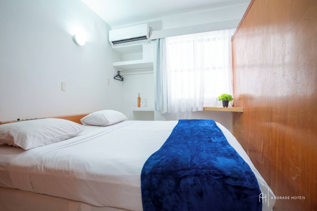 Quarto de hotel com cama, paredes brancas, aparelho de ar condicionado, janela com cortinas, pequena estante com planta e guarda-roupa de madeira.