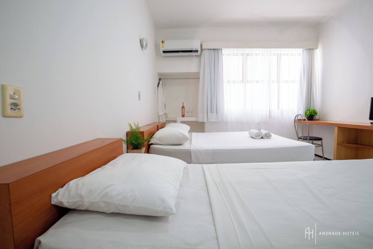 Quarto de hotel com duas camas, uma escrivaninha, uma cadeira, uma planta em vaso e uma janela com cortinas.