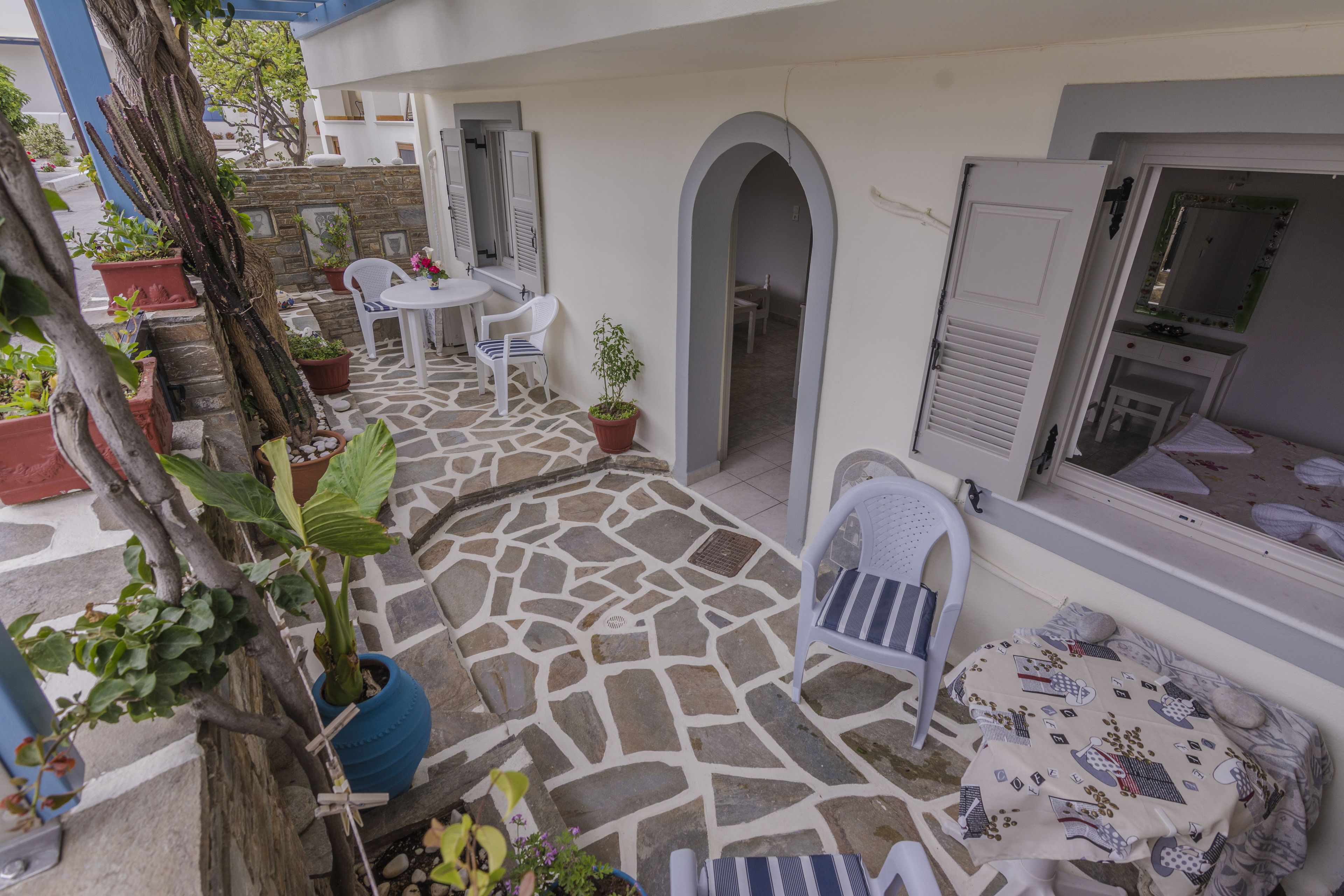 Un patio con pavimento in pietra, arredi bianchi, piante in vaso e un arco che conduce a una stanza con una toeletta e uno specchio.