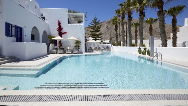 A swimming pool with lounge chairs and umbrellas, surrounded by white buildings and palm trees. - Kouros Village Hotel - Adults Only (Santorini)