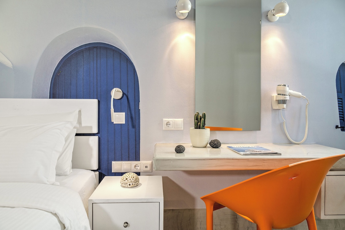 A hotel room with a bed, a desk with a chair, a mirror, a wall-mounted phone, and a potted plant.
