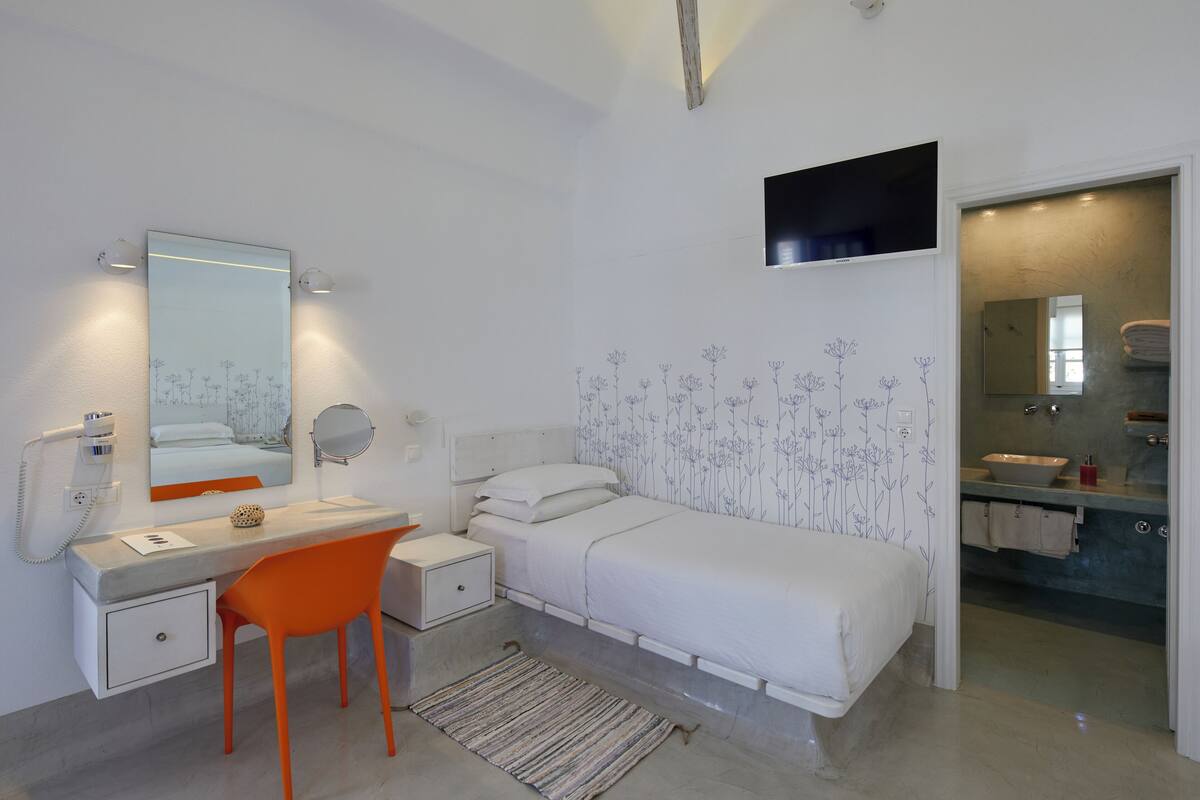 Un dormitorio con cama, escritorio, silla, mesita de noche, espejo y una ventana pequeña.
