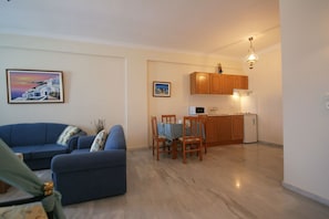 A living room with a sofa and a dining area with a table and chairs. There is a kitchen with wooden cabinets and a microwave. - Aegean View Hotel (Santorini)