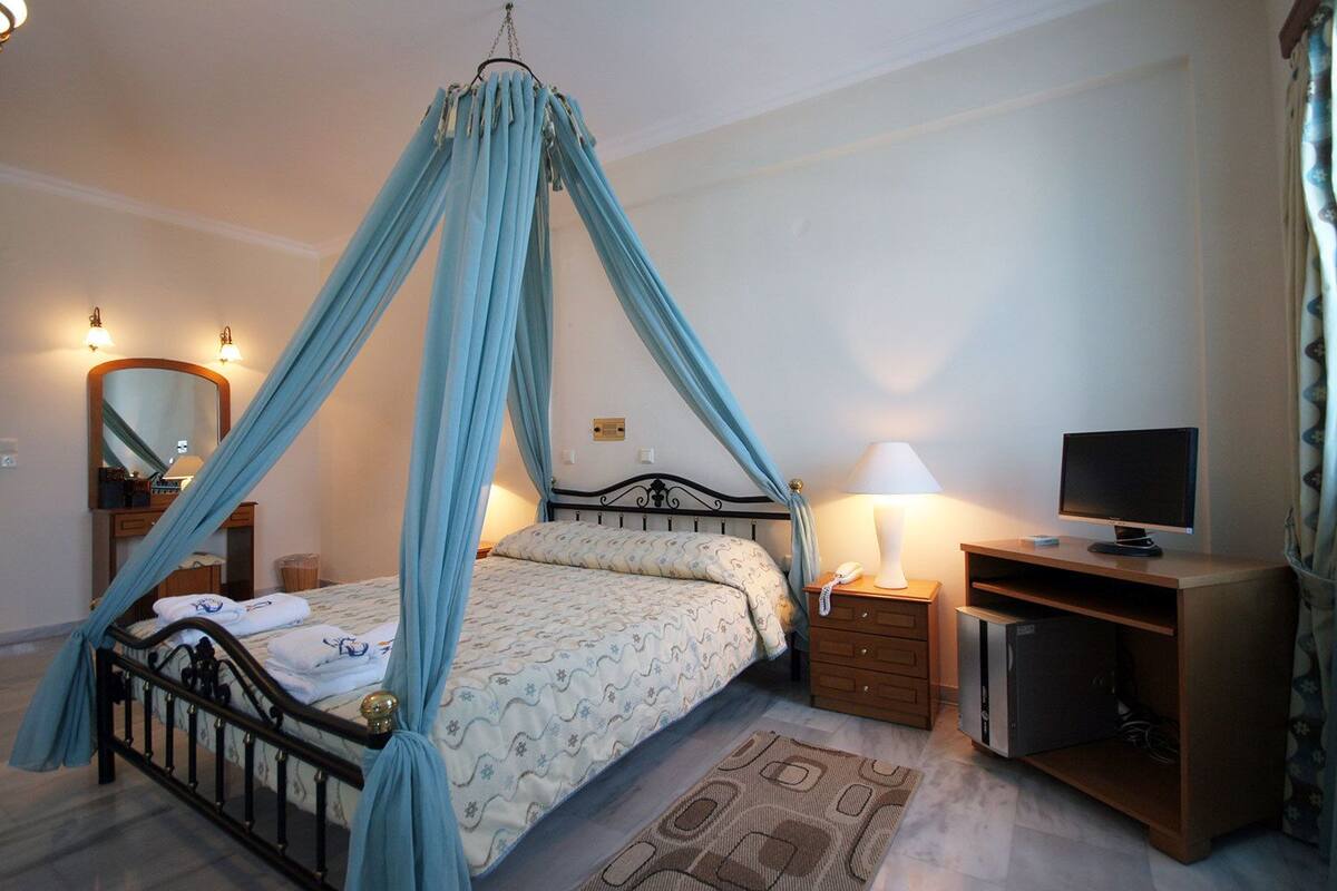 Ein Schlafzimmer mit einem Himmelbett, einem Nachttisch mit Lampe, einem Fernseher und einem kleinen Schreibtisch mit Stuhl.