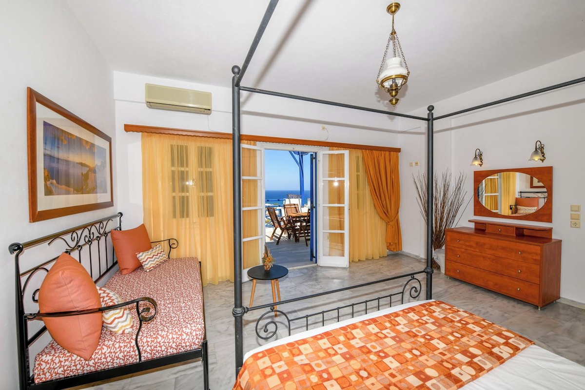 A bedroom with a four-poster bed, a dresser, a mirror, and a view of the sea through an open door.
