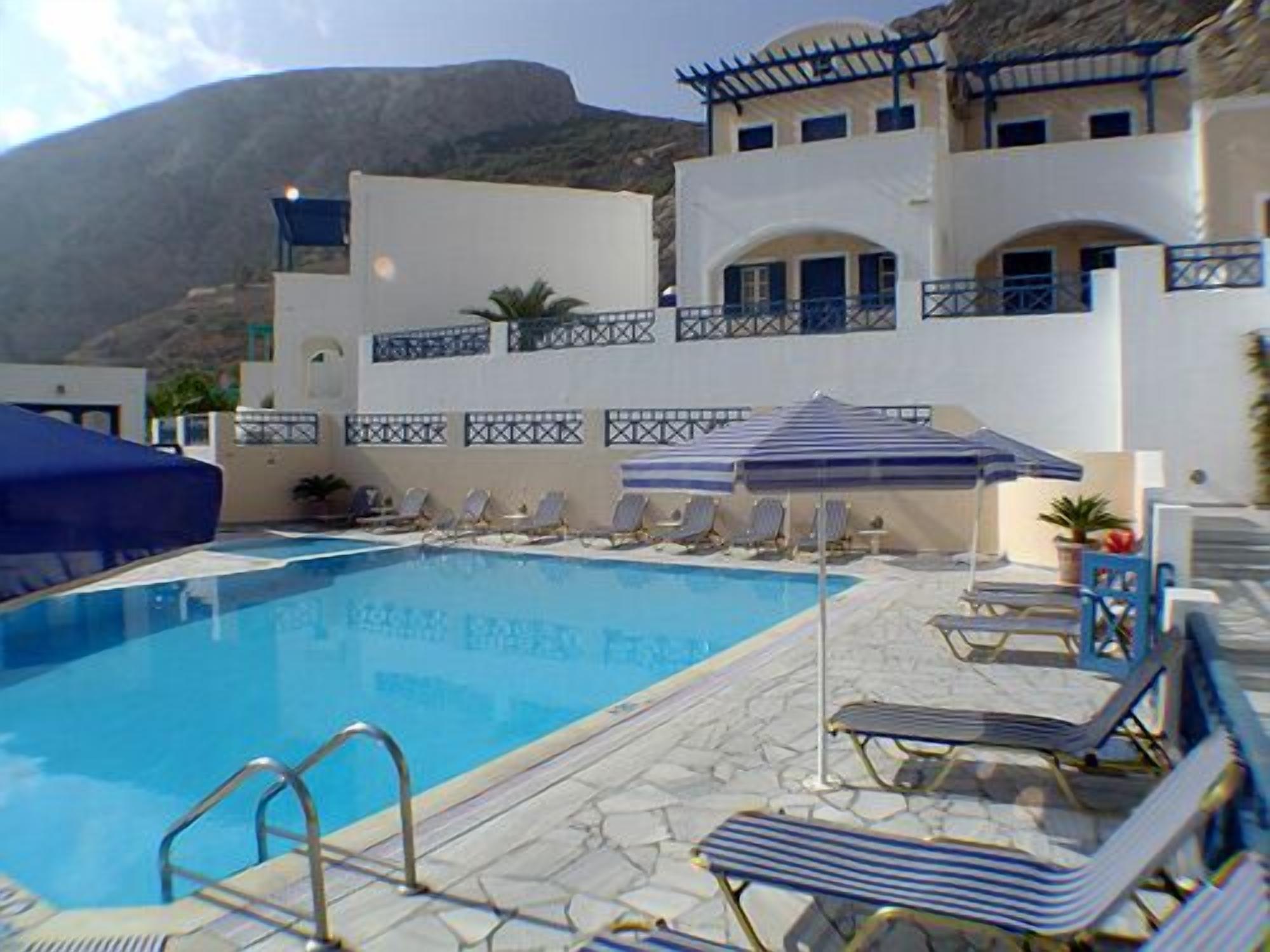 A pool area with lounge chairs and umbrellas, a clear blue pool, and a building with white walls and blue accents.