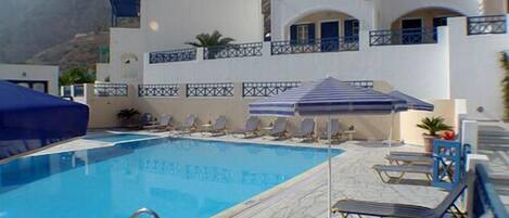 A pool area with lounge chairs and umbrellas, a clear blue pool, and a building with white walls and blue accents.