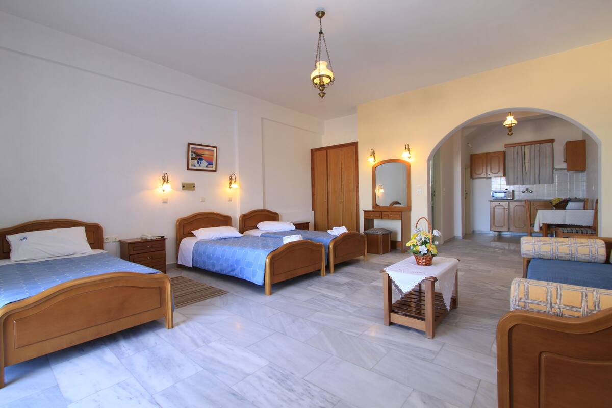 Ein Hotelzimmer mit drei Holzbett, einer Sitzecke mit Sofa und Sesseln, einem kleinen Tisch mit Korb und einer durch einen Bogen sichtbaren Küche.
