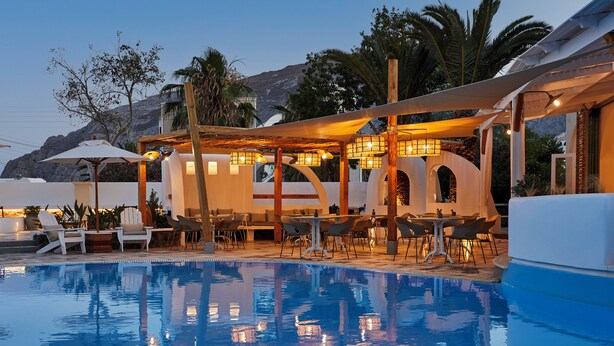 A pool area with lounge chairs and tables, surrounded by a covered seating area with lights. - Hermes Hotel (Santorini)