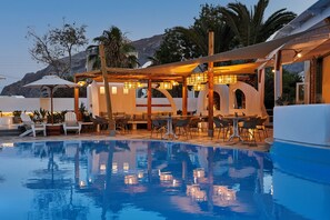 Un área de piscina con sillones y mesas, rodeada por una zona de estar cubierta con iluminación.