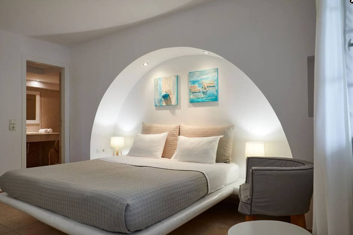 A bedroom with a bed, two wall-mounted lamps, a chair, and two paintings.