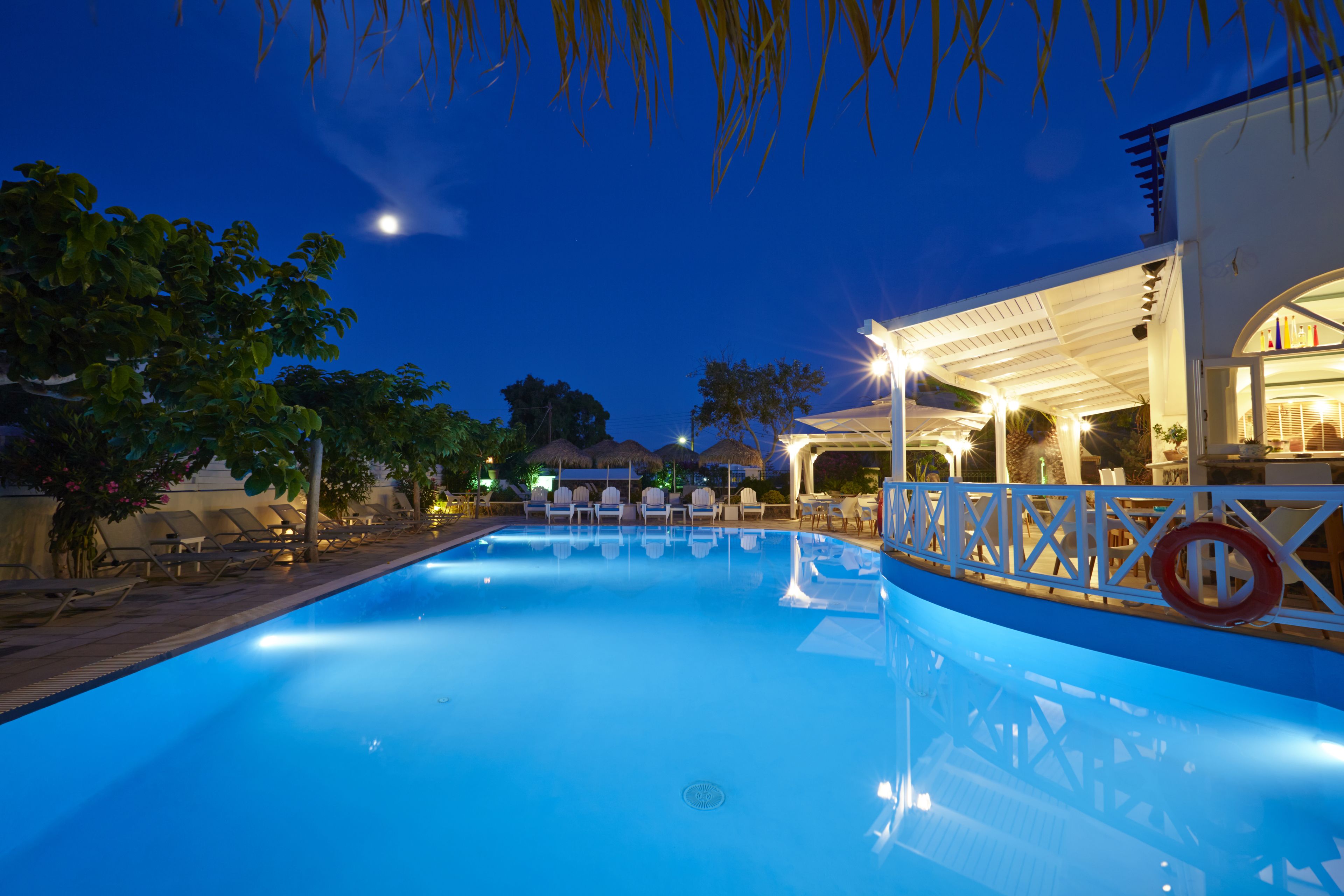 A swimming pool with lounge chairs and umbrellas, surrounded by trees and a building with a covered patio.