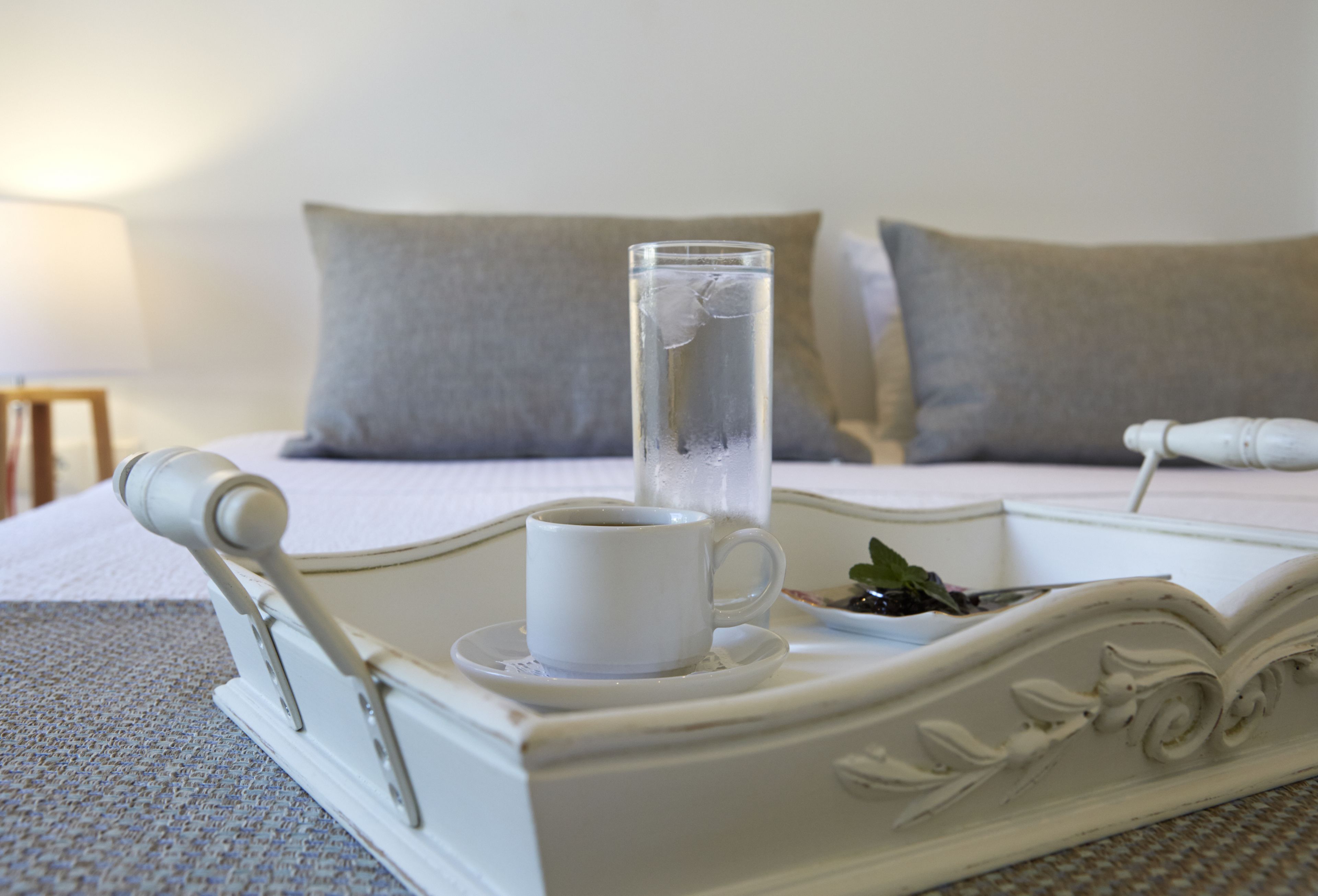 Una bandeja blanca con una taza de café y un vaso de agua sobre una cama con almohadillas grises.